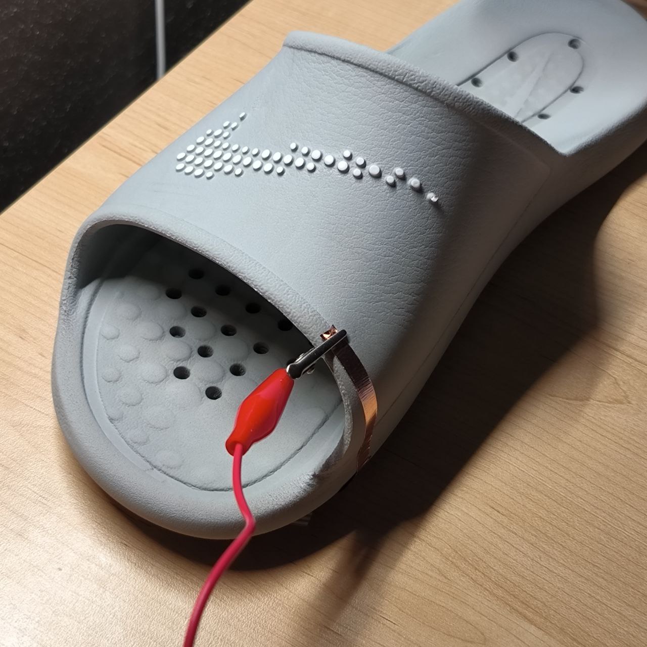 A clip connects an alligator wire to the side of the slipper, suggesting the start of testing conductivity or creating a touchpoint. This is the first step in transforming the slipper into an interactive controller—potentially usable with a system like Makey Makey.
