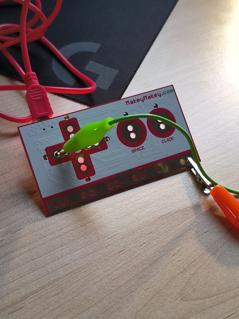 A Makey Makey board is powered and ready, with alligator clips attached. One clip is connected to the space input, preparing the board to detect touch-based interactions from the paper circuit.